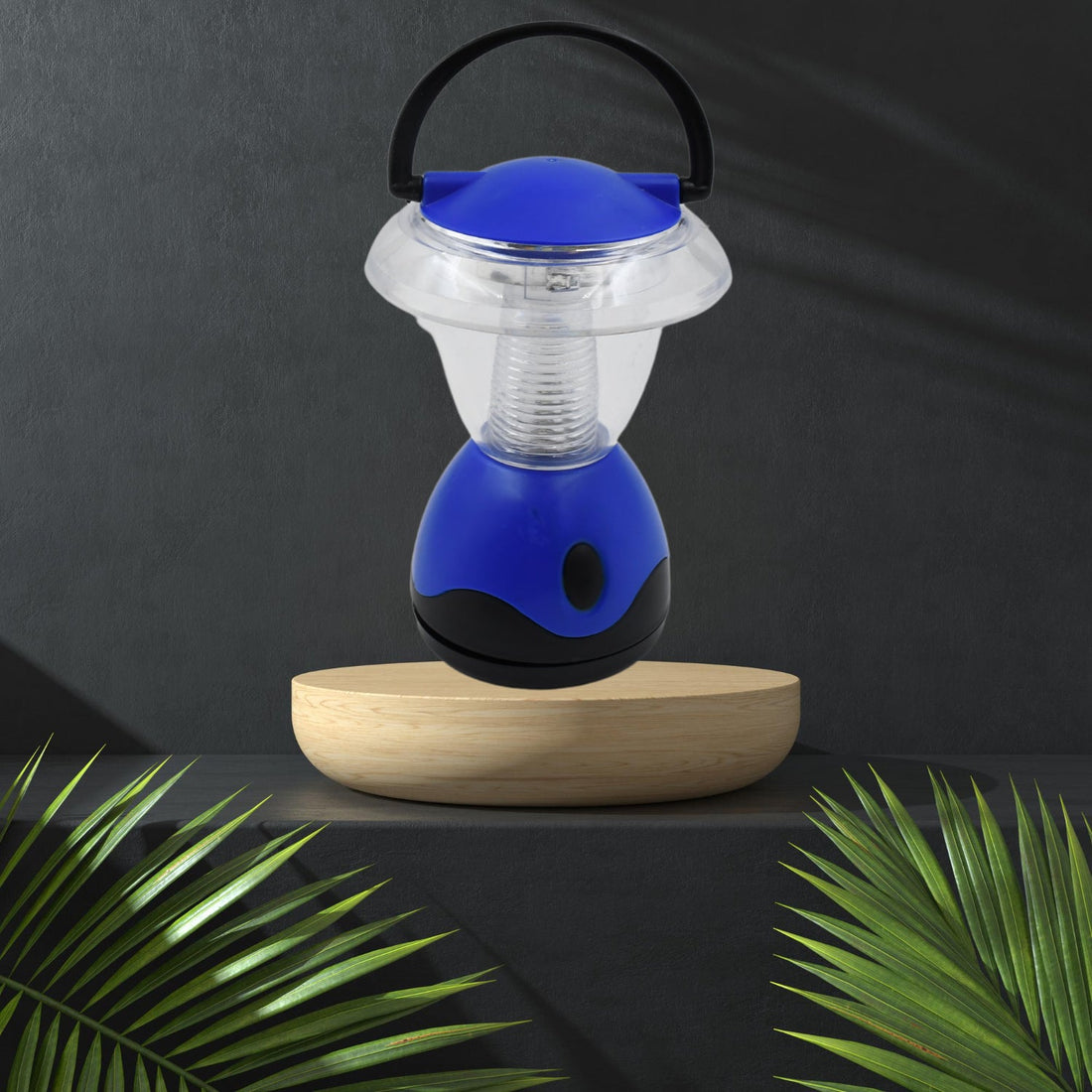 The blue and clear plastic mini lantern rests on a light wooden pedestal, flanked by green palm fronds against a dark